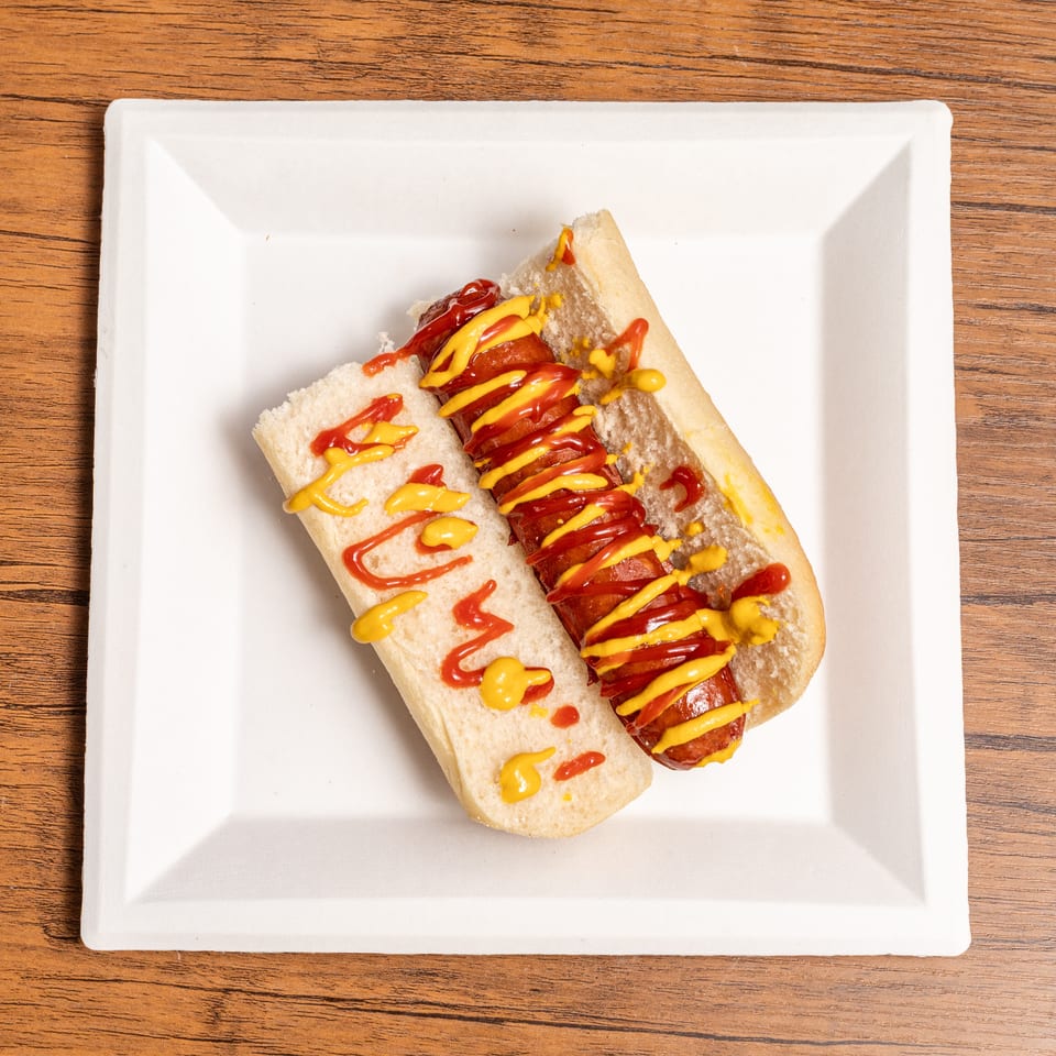 Hot dog with ketchup and mustard on a white plate, on a wooden table.