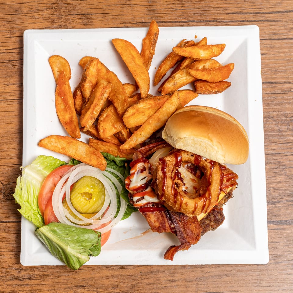 A burger with bacon, cheese, onion rings, and sauces, served with potato wedges and a side salad.