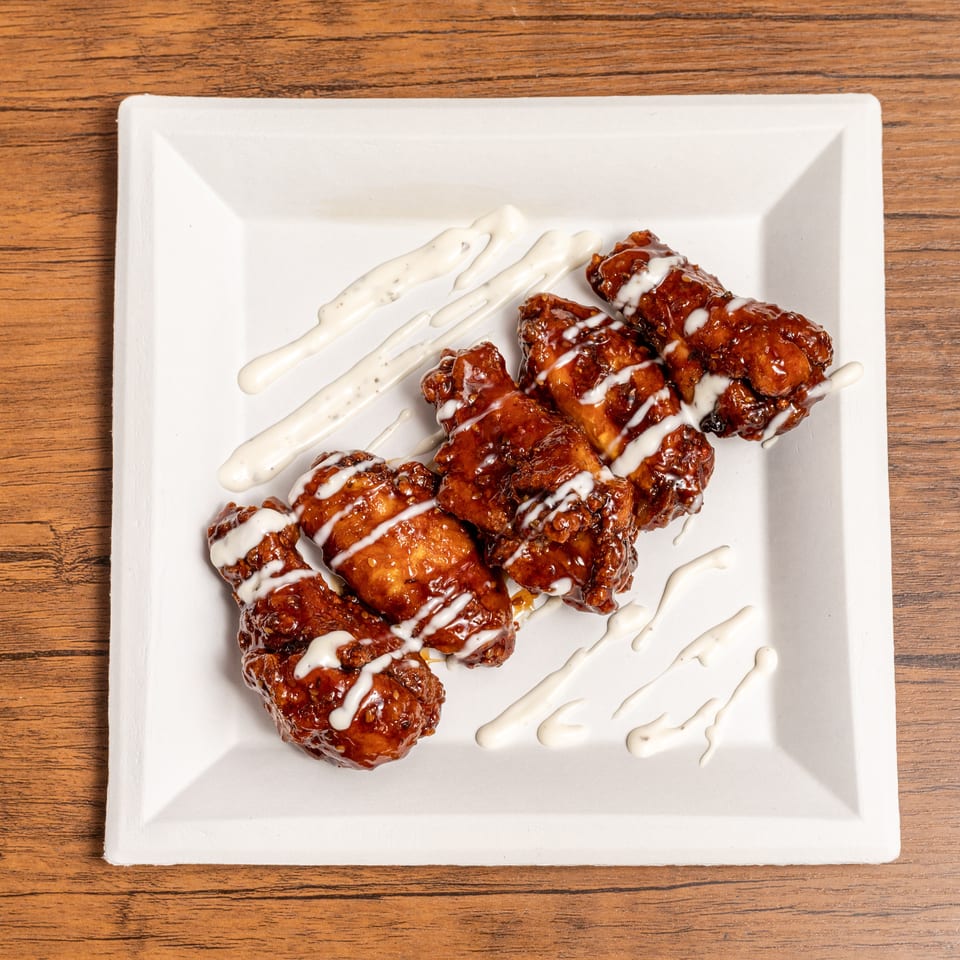 Glazed fried chicken pieces drizzled with creamy white sauce on a square white plate atop a wooden table.