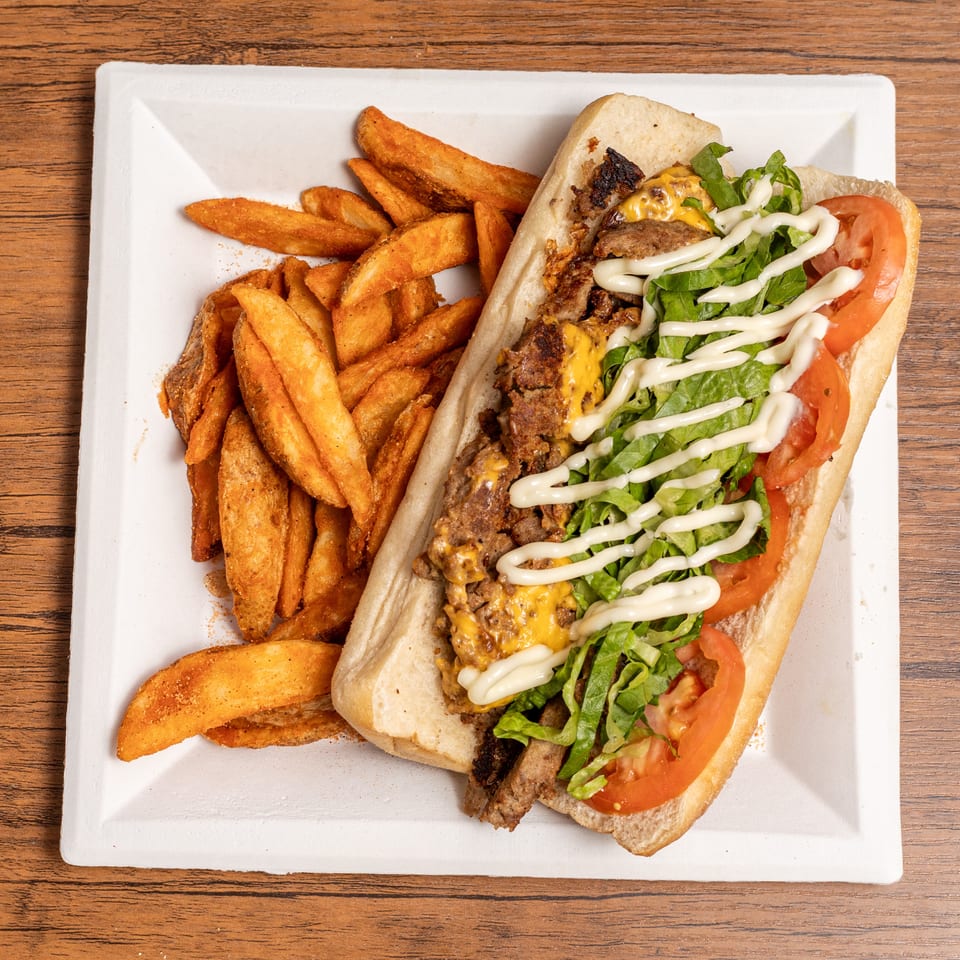 Cheesesteak sandwich with lettuce, tomato, and mayonnaise, served with seasoned potato wedges on a white plate.