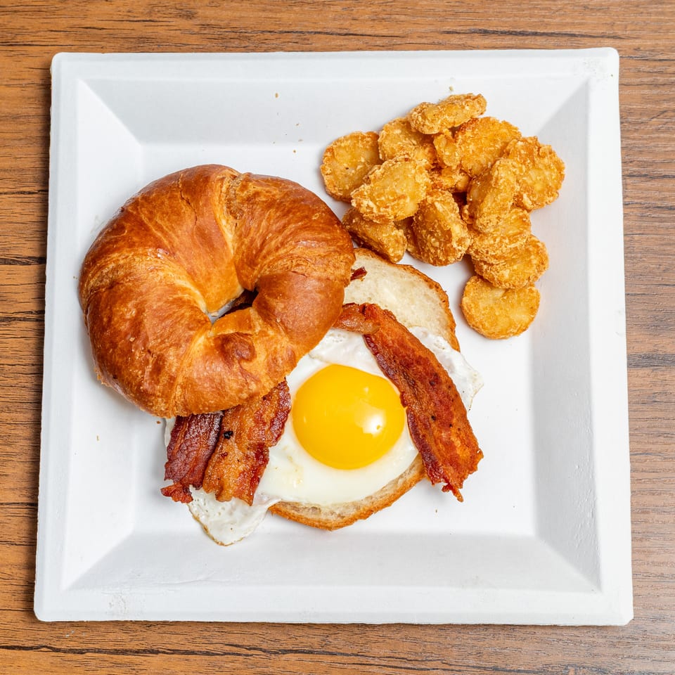 Breakfast plate with a croissant, fried egg, bacon, and hash browns on a white plate.
